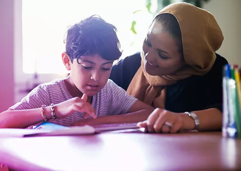 A young person sitting at a table with colouring pencils and paper gets encouragement from a tutor.